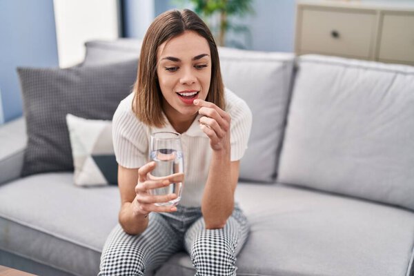 Young beautiful hispanic woman taking pill sitting on sofa at home
