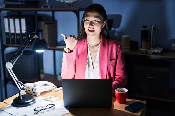 Chinese young woman working at the office at night smiling with happy face looking and pointing to the side with thumb up.