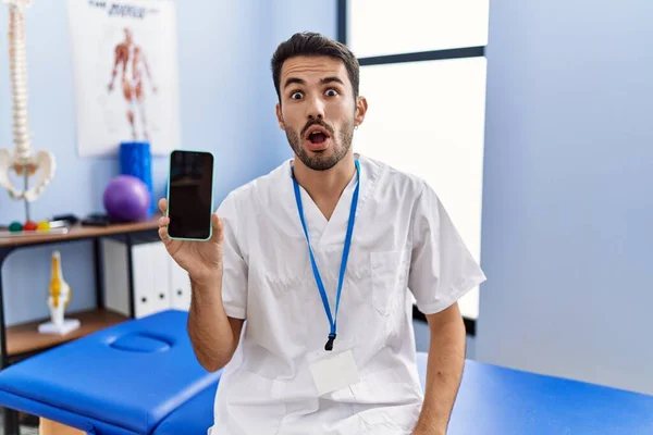 Young Hispanic Physiotherapist Man Holding Smartphone Clinic Scared Amazed Open — Stockfoto