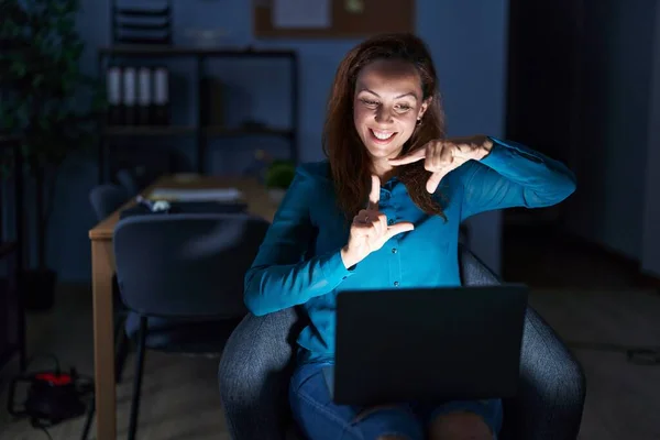 Brunette woman working at the office at night smiling making frame with hands and fingers with happy face. creativity and photography concept.