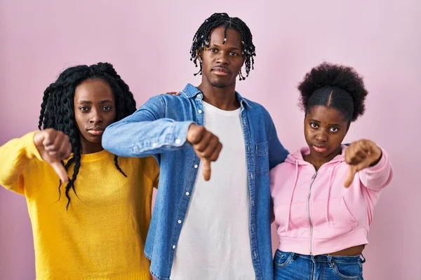 Group of three young black people standing together over pink background looking unhappy and angry showing rejection and negative with thumbs down gesture. bad expression.