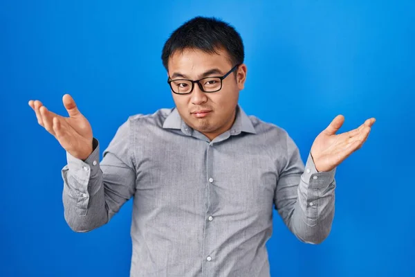 Young chinese man standing over blue background clueless and confused expression with arms and hands raised. doubt concept.