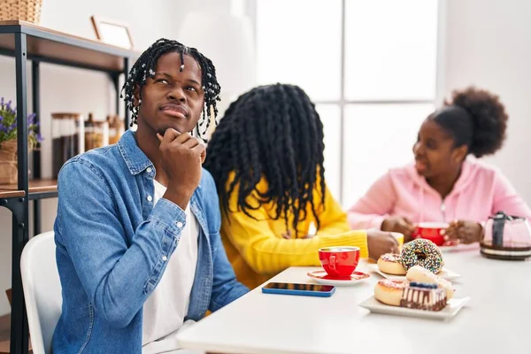 Group of three young black people sitting on a table having coffee serious face thinking about question with hand on chin, thoughtful about confusing idea