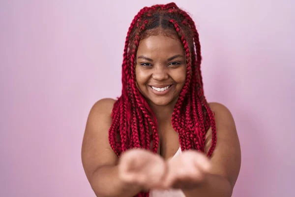 African american woman with braided hair standing over pink background smiling with hands palms together receiving or giving gesture. hold and protection