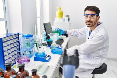 Young hispanic man wearing scientist uniform pouring liquid on test bottle at laboratory