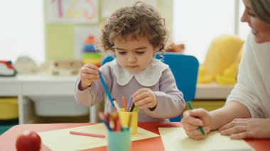 Teacher and toddler sitting on table drawing on paper at kindergarten