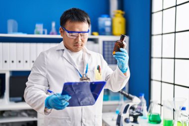 Young chinese man wearing scientist uniform reading clipboard at laboratory