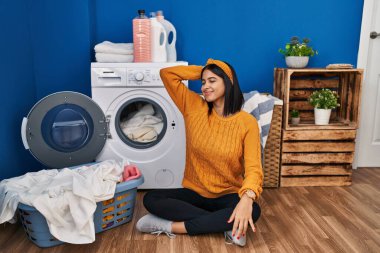 Young hispanic woman doing laundry smiling confident touching hair with hand up gesture, posing attractive and fashionable 