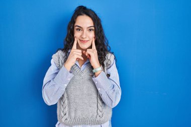 Genç esmer kadın mavi arka planda duruyor ağzı açık gülümsüyor, parmakları işaret ediyor ve zorla neşeli bir gülümseme sergiliyor. 