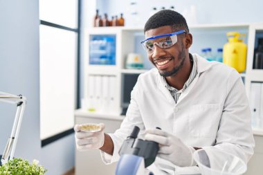 Genç Afrikalı Amerikalı adam laboratuarda mikroskop kullanarak bilim adamı üniforması giyiyor.