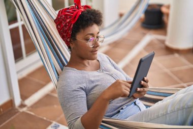 Ev terasında hamakta uzanmış dokunmatik ped kullanan Afrikalı Amerikalı bir kadın.