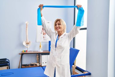 Middle age blonde woman wearing physiotherapist uniform using elastic band at physiotherapy clinic