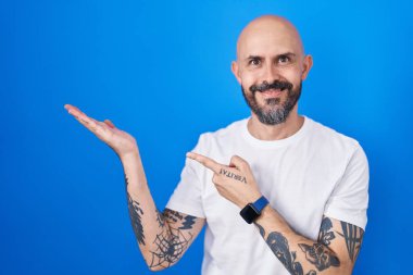 Hispanic man with tattoos standing over blue background amazed and smiling to the camera while presenting with hand and pointing with finger. 