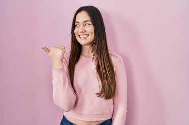 Young brunette woman standing over pink background smiling with happy face looking and pointing to the side with thumb up. 