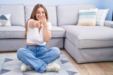 Young caucasian woman sitting on the floor at the living room laughing at you, pointing finger to the camera with hand over mouth, shame expression 