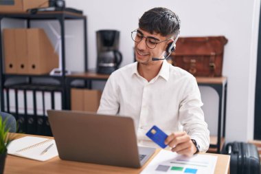 Young hispanic man working using computer laptop holding credit card looking away to side with smile on face, natural expression. laughing confident. 