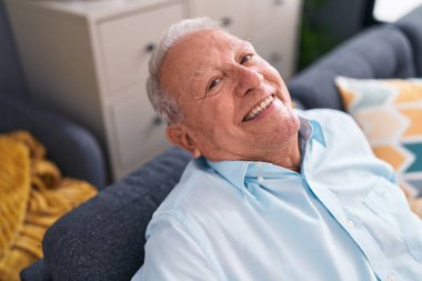 Orta yaşlı, kır saçlı, gülümseyen bir adam. Evdeki kanepede oturuyor.