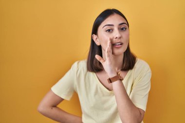 Hispanic girl wearing casual t shirt over yellow background hand on mouth telling secret rumor, whispering malicious talk conversation 