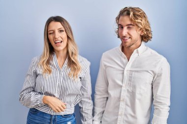 Young couple standing over blue background winking looking at the camera with sexy expression, cheerful and happy face. 