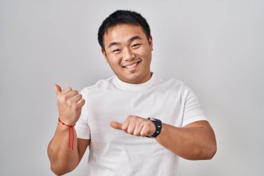 Young chinese man standing over white background pointing to the back behind with hand and thumbs up, smiling confident 