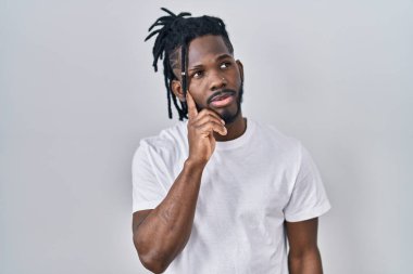 Rasta saçlı Afrikalı bir adam beyaz arka plan üzerine gündelik tişört giyiyor, elleri çenesinde soru sorarken, düşünceli bir ifade takınıyor. Düşünceli bir yüzle gülümsüyor. şüphe kavramı. 