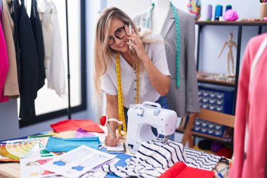 Terzi dükkanında akıllı telefondan konuşan genç sarışın kadın deftere yazı yazıyor.