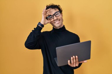 Hispanic man working using computer laptop smiling happy doing ok sign with hand on eye looking through fingers 