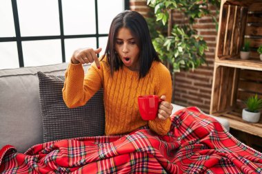 Young hispanic woman sitting on the sofa drinking a coffee at home pointing down with fingers showing advertisement, surprised face and open mouth 