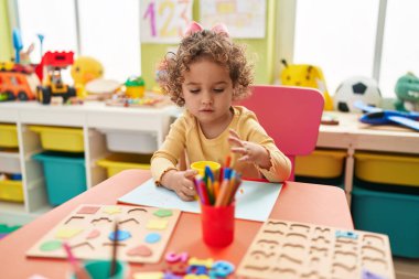 Adorable hispanic toddler student sitting on table drawing on paper at kindergarten