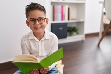 Adorable hispanic boy student smiling confident reading book at library school