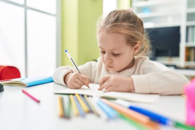 Adorable blonde girl student writing on notebook sitting on table at classroom