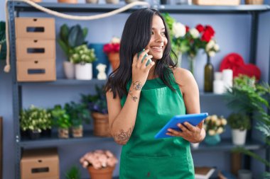 Young hispanic woman florist talking on smartphone using touchpad at florist shop