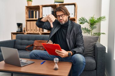 Handsome middle age man working at consultation office looking unhappy and angry showing rejection and negative with thumbs down gesture. bad expression. 