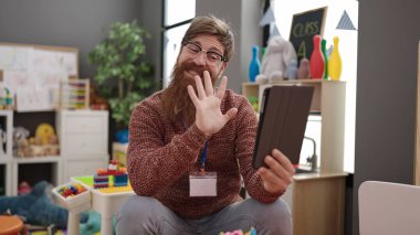 Young redhead man preschool teacher having video call sitting on chair at kindergarten
