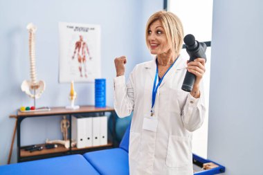 Middle age blonde woman holding therapy massage gun at physiotherapy center pointing thumb up to the side smiling happy with open mouth 