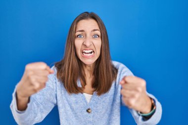 Young woman standing over blue background angry and mad raising fists frustrated and furious while shouting with anger. rage and aggressive concept. 