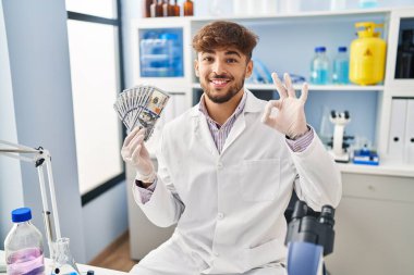 Arab man with beard working at scientist laboratory holding money doing ok sign with fingers, smiling friendly gesturing excellent symbol 