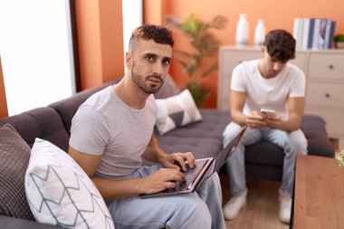 Two hispanic men couple using laptop and smartphone sitting on sofa at home
