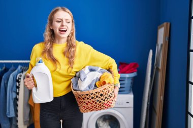 Young caucasian woman holding laundry basket and detergent bottle angry and mad screaming frustrated and furious, shouting with anger. rage and aggressive concept. 