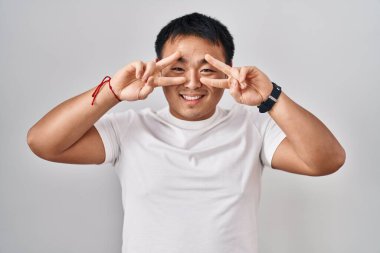 Young chinese man standing over white background doing peace symbol with fingers over face, smiling cheerful showing victory 