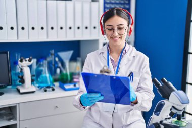 Young hispanic woman scientist listening to music writing on document at laboratory