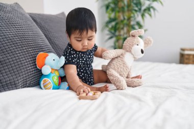 Adorable hispanic baby playing with dolls sitting on bed at bedroom