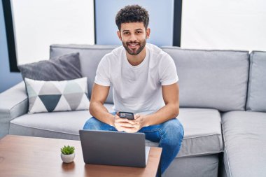 Young arab man using smartphone and laptop sitting on sofa at home