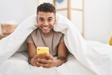Afrika kökenli Amerikalı bir adam yatakta akıllı telefon kullanıyor.