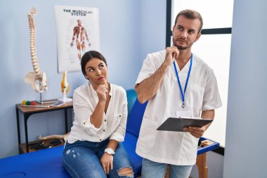 Young hispanic woman at physiotherapist appointment with hand on chin thinking about question, pensive expression. smiling with thoughtful face. doubt concept. 