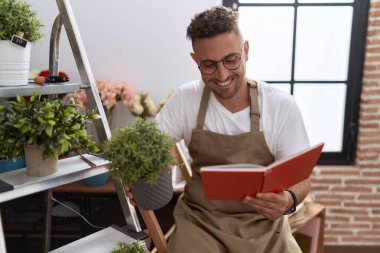 Young hispanic man florist reading notebook holding plant at flower shop