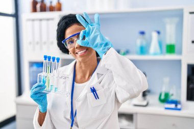 Middle age hispanic woman working at scientist laboratory smiling happy doing ok sign with hand on eye looking through fingers 