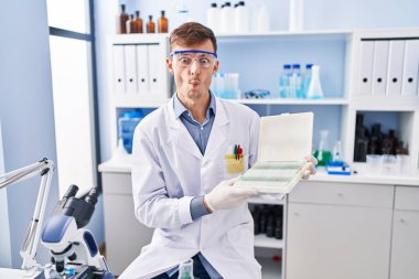Caucasian man working at scientist laboratory making fish face with mouth and squinting eyes, crazy and comical. 