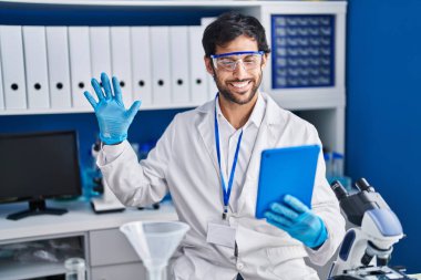 Handsome latin man working at scientist laboratory using tablet looking positive and happy standing and smiling with a confident smile showing teeth 