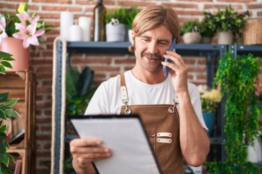 Çiçekçide akıllı telefondan konuşan genç sarışın adam pano okuyor.